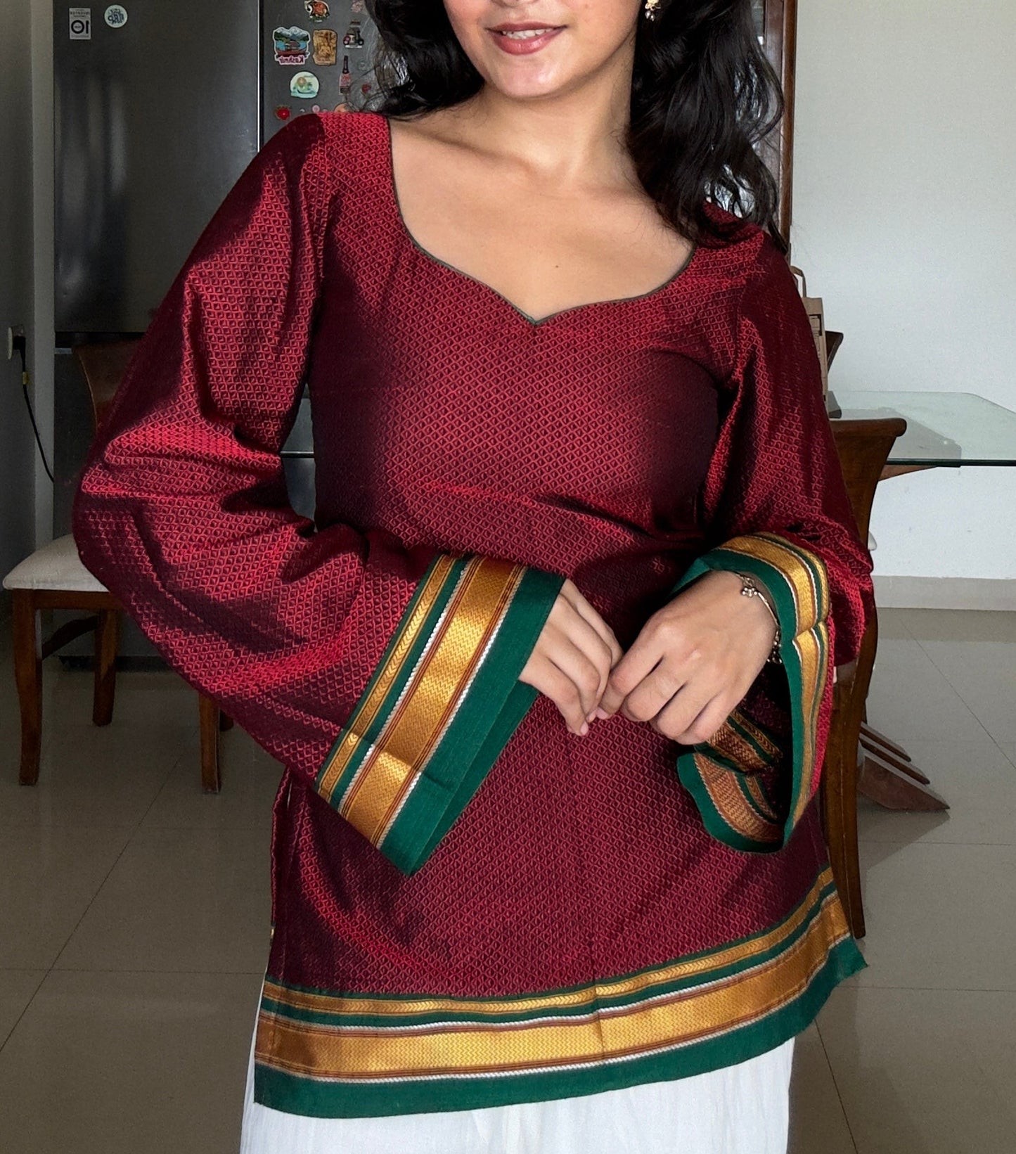 A woman wearing a maroon long-sleeve khunn kurta with green and gold striped cuffs and hem, paired with white pants, standing in a home interior.