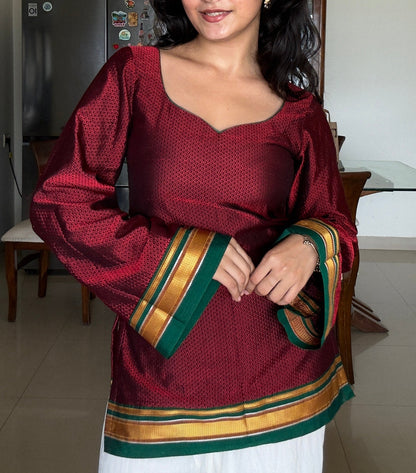 A woman wearing a maroon long-sleeve khunn kurta with green and gold striped cuffs and hem, paired with white pants, standing in a home interior.