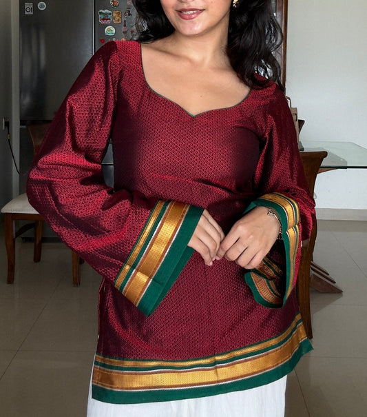 A woman wearing a maroon long-sleeve khunn kurta with green and gold striped cuffs and hem, paired with white pants, standing in a home interior.