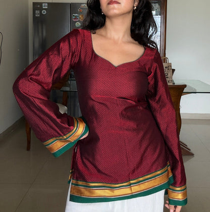 A woman wearing a traditional long-sleeve kurta in deep maroon with a subtle geometric pattern, featuring a green and gold striped border at the hem and cuffs, paired with white trousers in an indoor home setting.